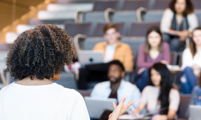 A photo of a female professor in a lecture hall teaching a group of students. Une photo d'une professeure dans une salle de conférence enseignant à un groupe d'étudiant.e.s.