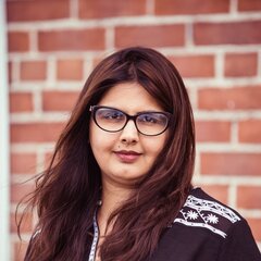 Photo of Shumaila Hemani looking at the camera and smiling in front of a brick wall.